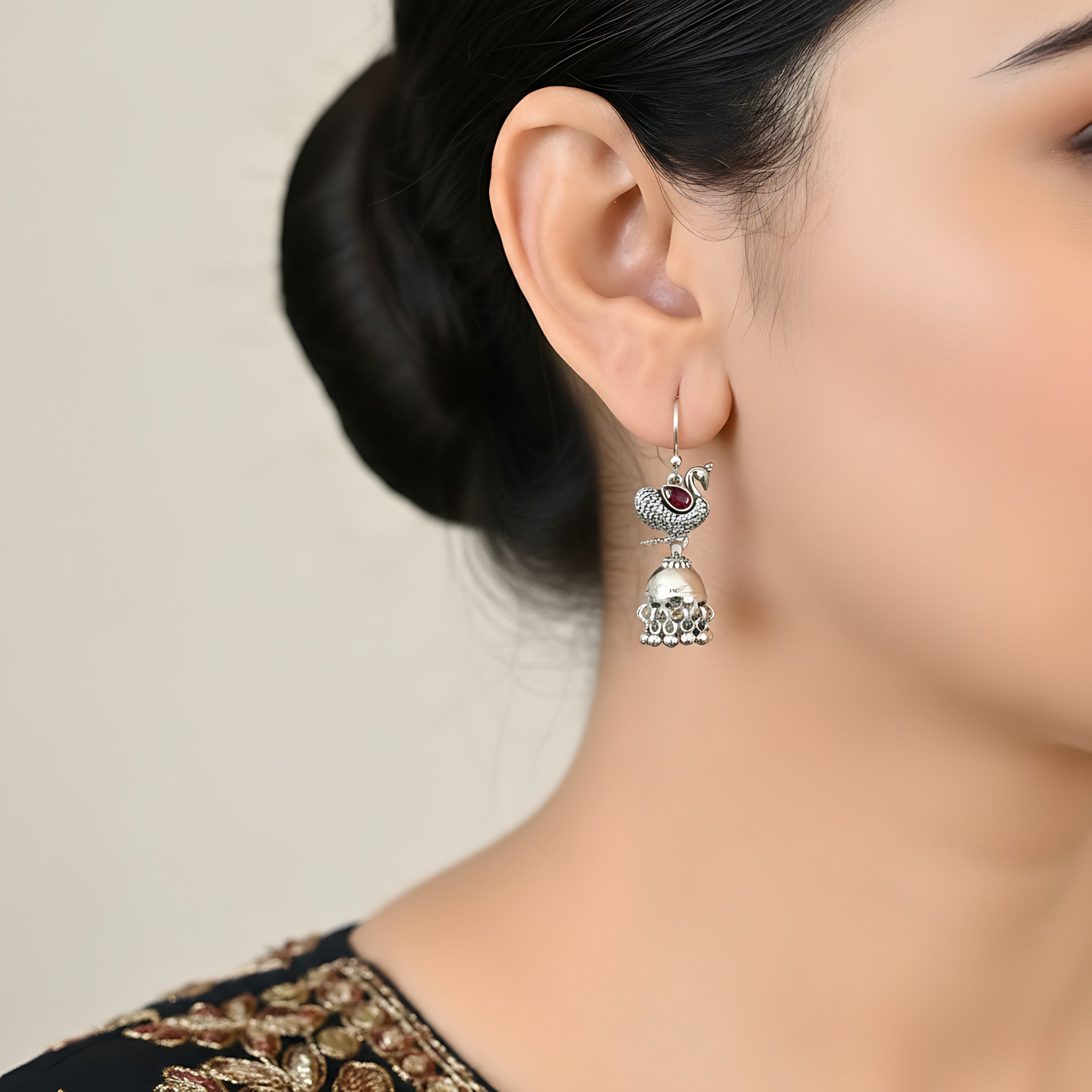 Close-up of a woman wearing an oxidized silver peacock jhumka earring with a red gemstone on a neutral background