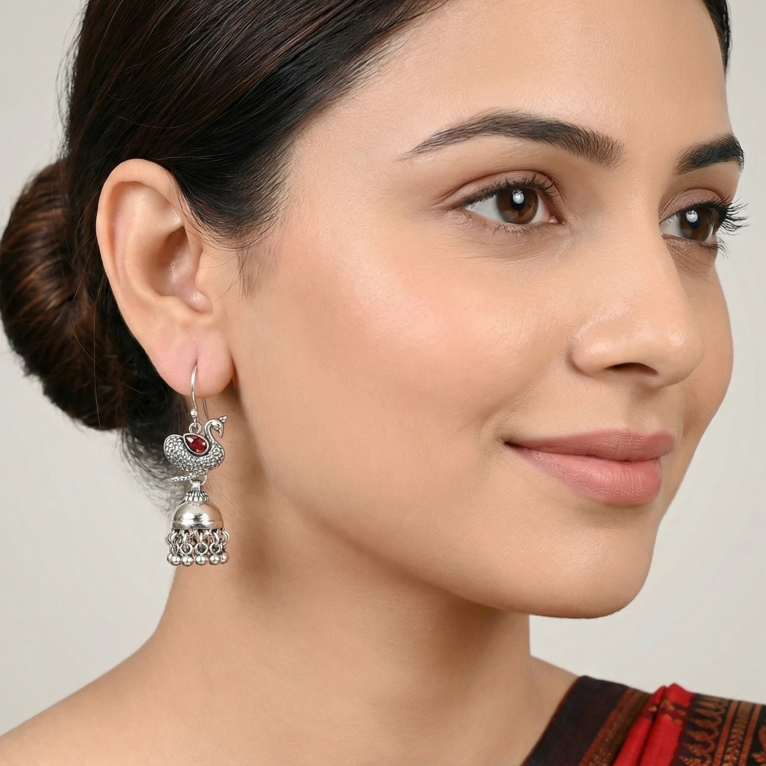 Close-up of a woman wearing an oxidized silver peacock jhumka earring with a red gemstone on a neutral background