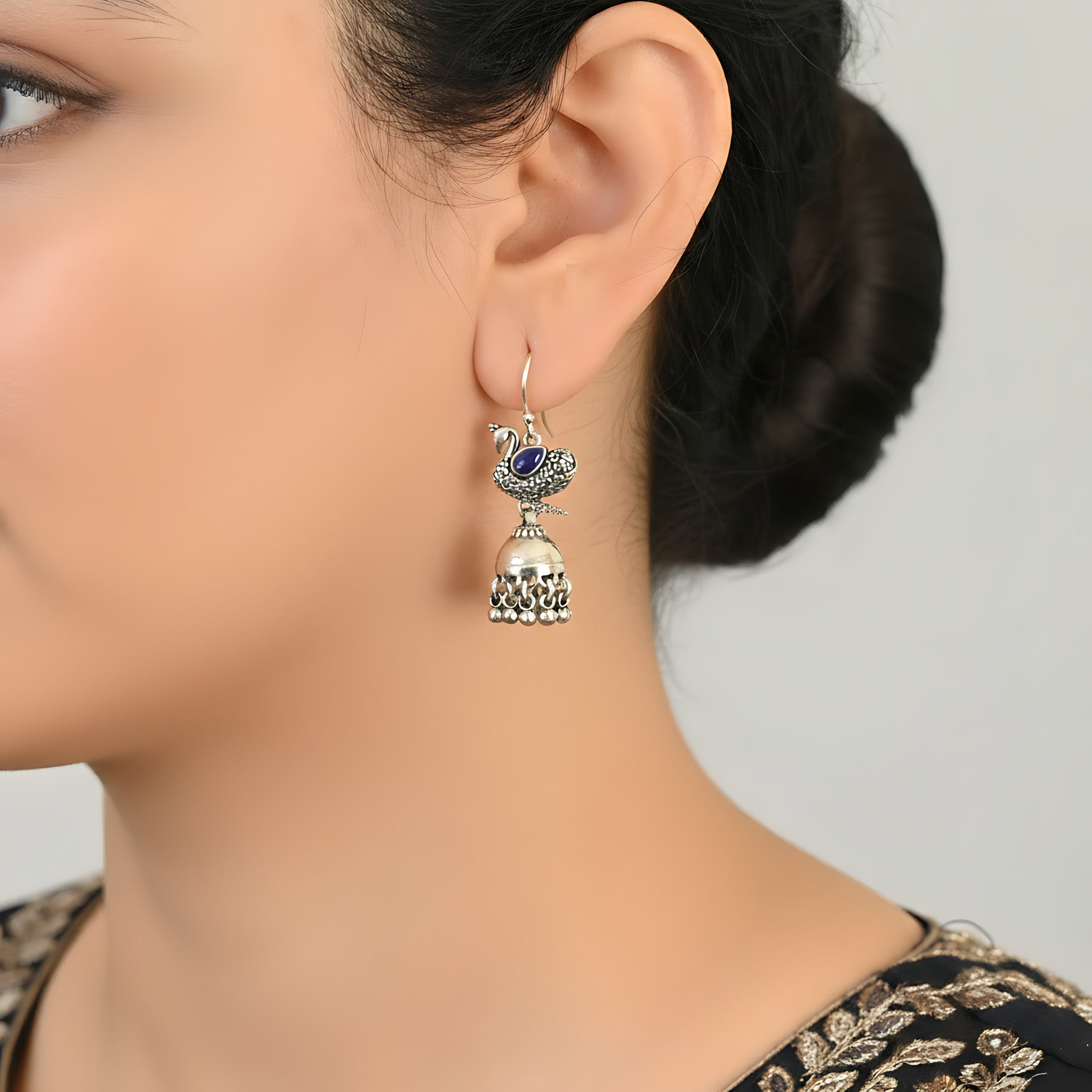 Close-up of a woman wearing an oxidized silver peacock jhumka earring with a blue gemstone on a neutral background