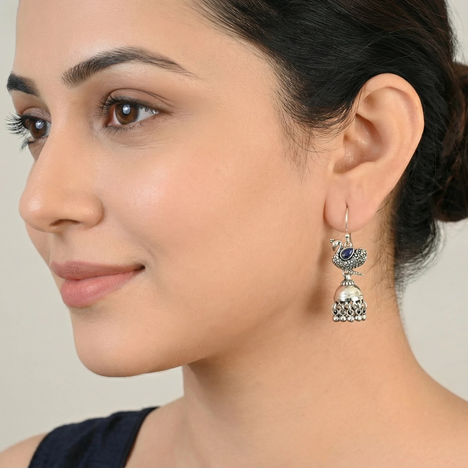 Close-up of a woman wearing an oxidized silver peacock jhumka earring with a blue gemstone on a neutral background