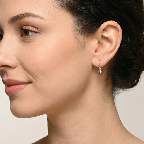 Close-up of a woman wearing a Silver Ball Charm Hoop Earring with a clear gemstone on a neutral background