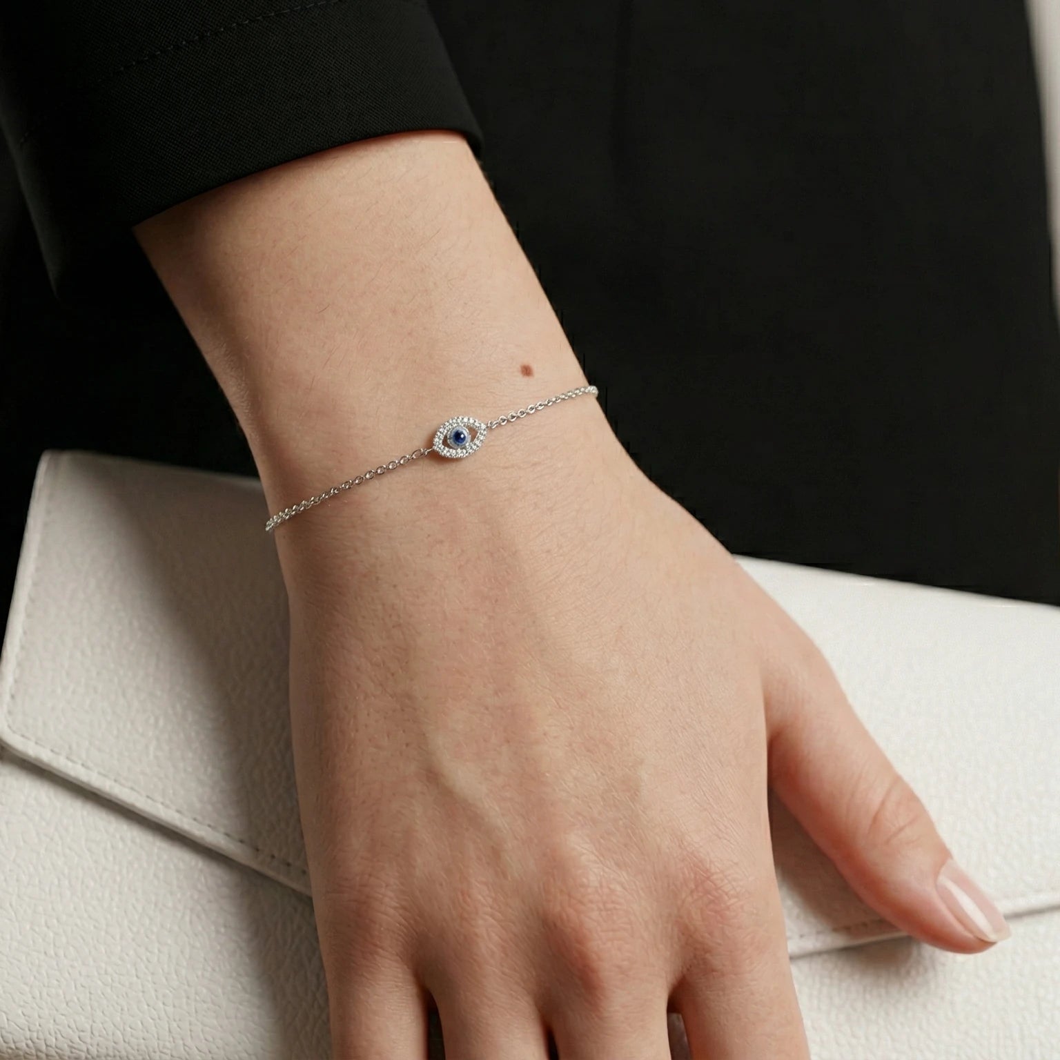 Hand wearing a silver evil eye bracelet, holding a white clutch against a dark background