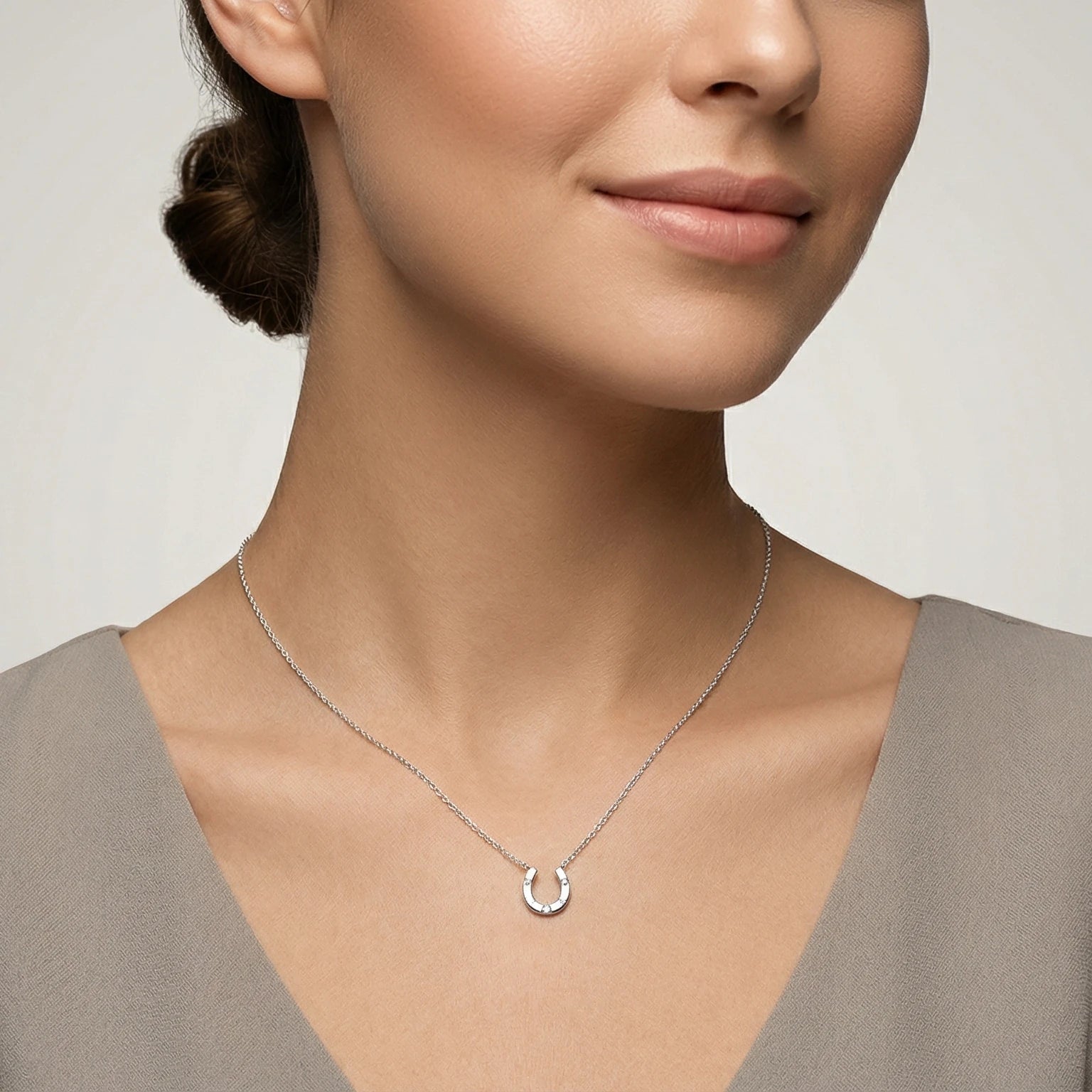 Woman wearing a delicate silver necklace with a small horseshoe pendant against a neutral background