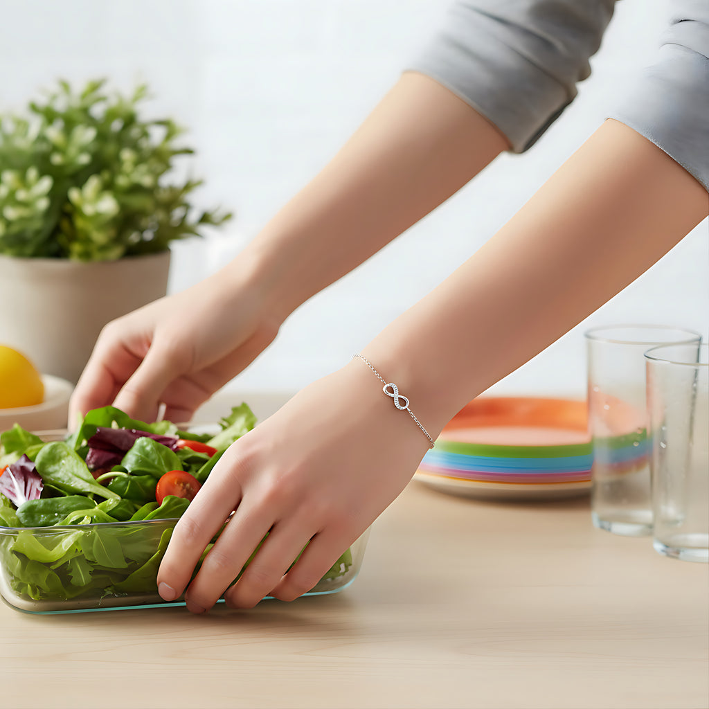 Person wearing a silver infinity bracelet preparing a salad  on a table with a plant and glasses in the background