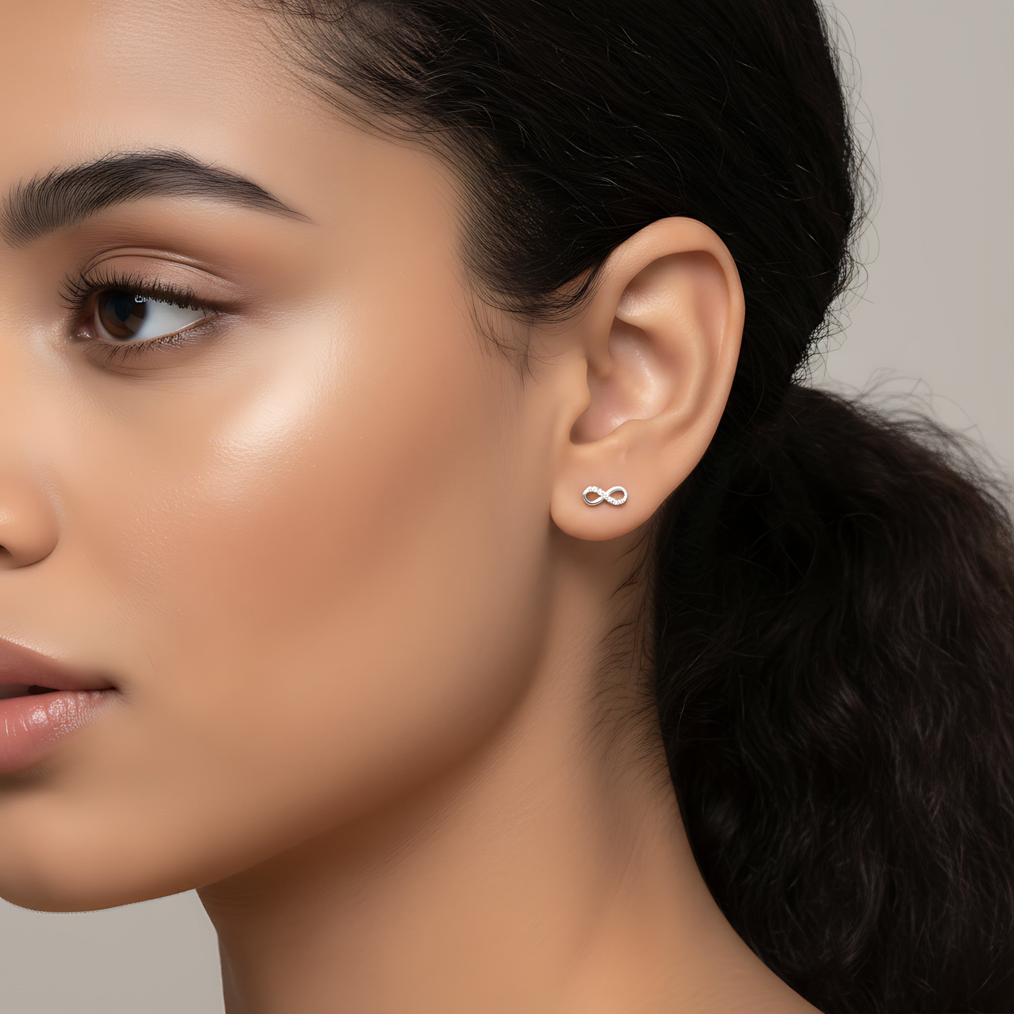 Woman with short dark hair wearing a delicate infinity shaped silver stud earring against a plain background