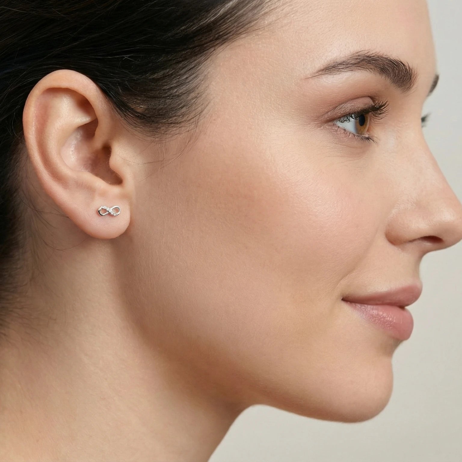 Woman wearing a delicate infinity shaped silver stud earring against a plain background