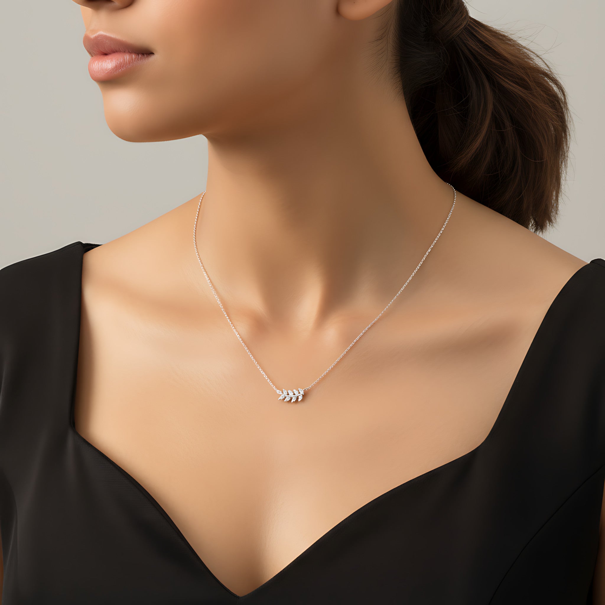 Closeup of a female wearing a silver necklace with a small olive leaf pendant against a neutral background