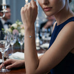 Woman wearing a delicate clover charm bracelet at a formal event with wine glasses and flowers in the background.