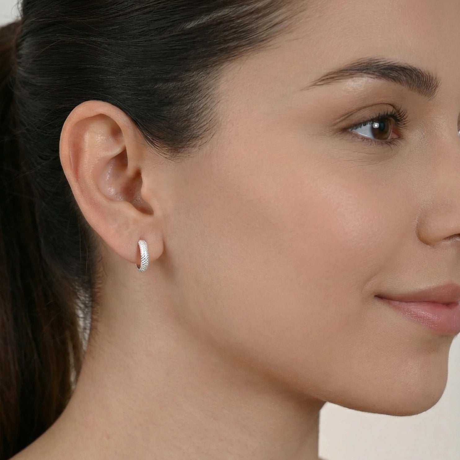 Silver hoop earring with diamond cut texture worn by a female with a neutral background