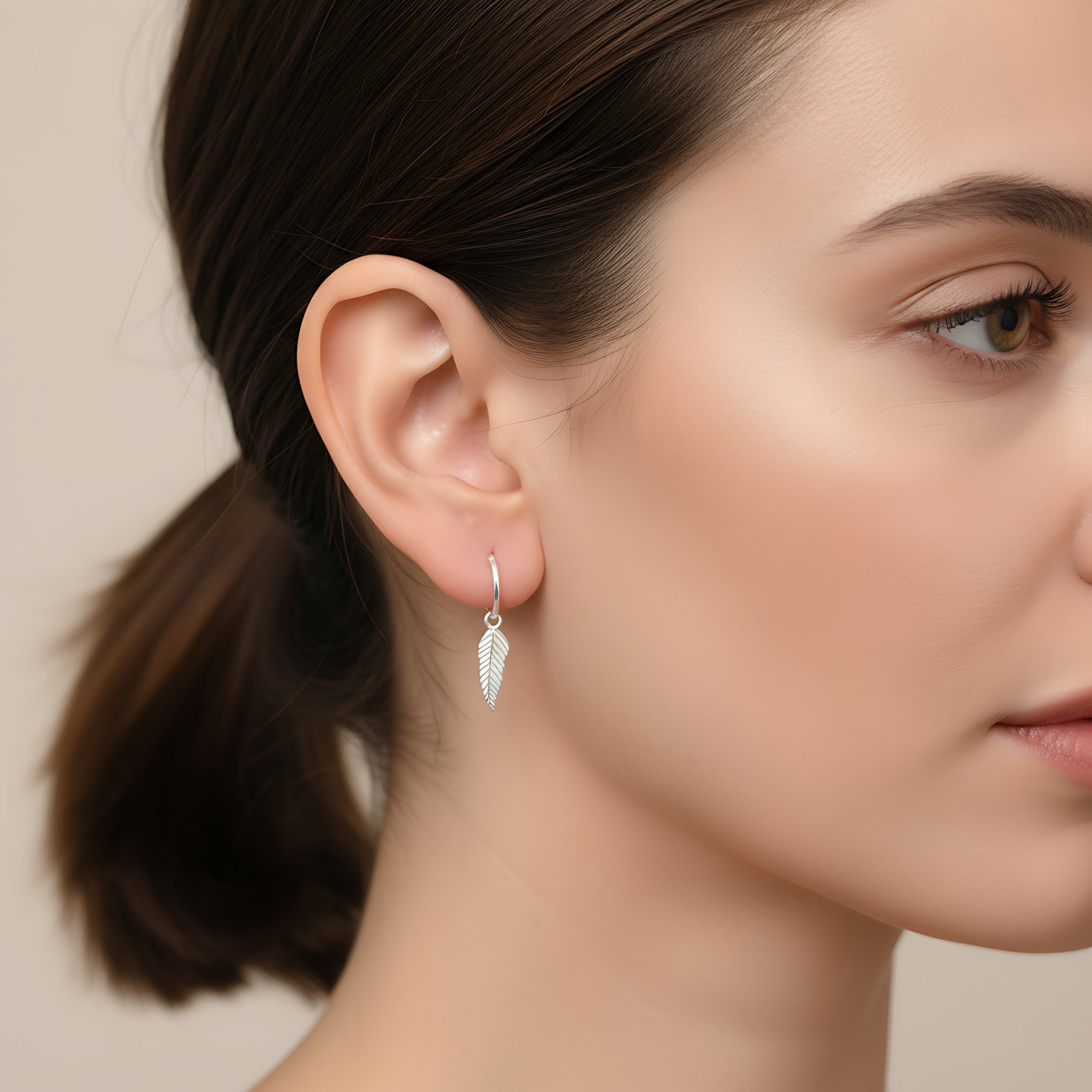 Close-up of a woman wearing silver feather charm hoop earring with a neutral background