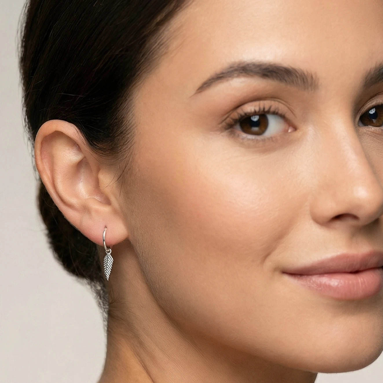 Close-up of a woman wearing silver feather charm hoop earring with a neutral background