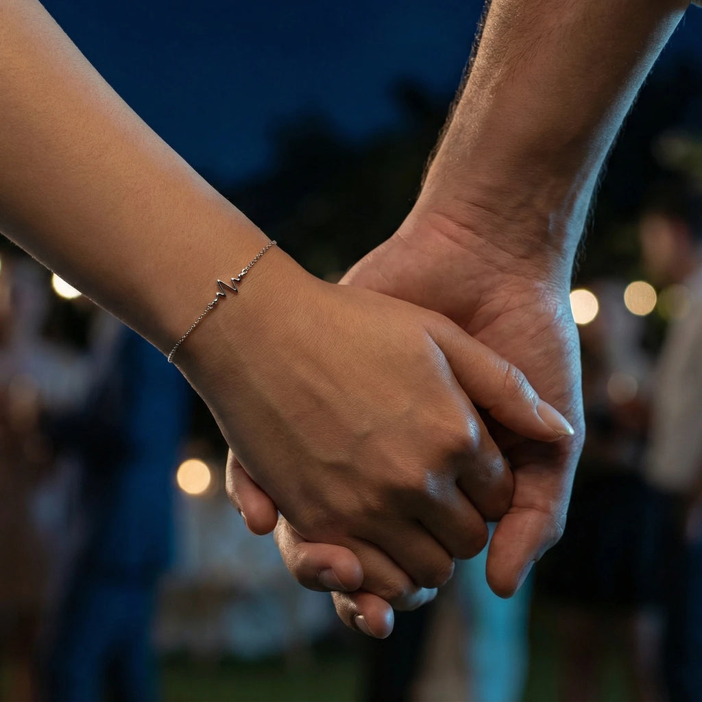 Two hands holding each other with a blurred background of lights and the female is wearing a heartbeat charm silver bracelet.