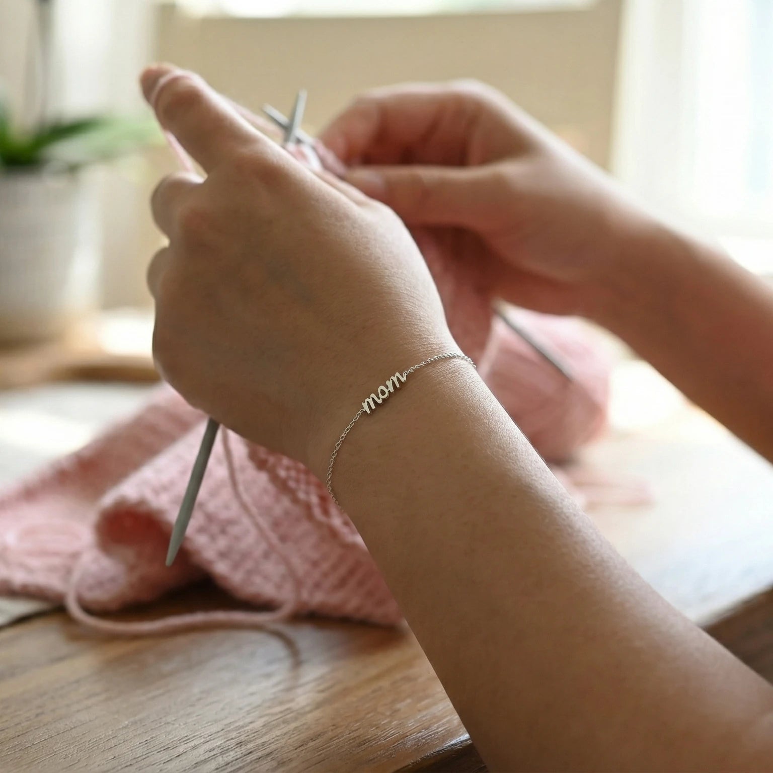 Close-up of hands knitting with yarn and needles on a wooden surface and wearing a mom silver bracelet.