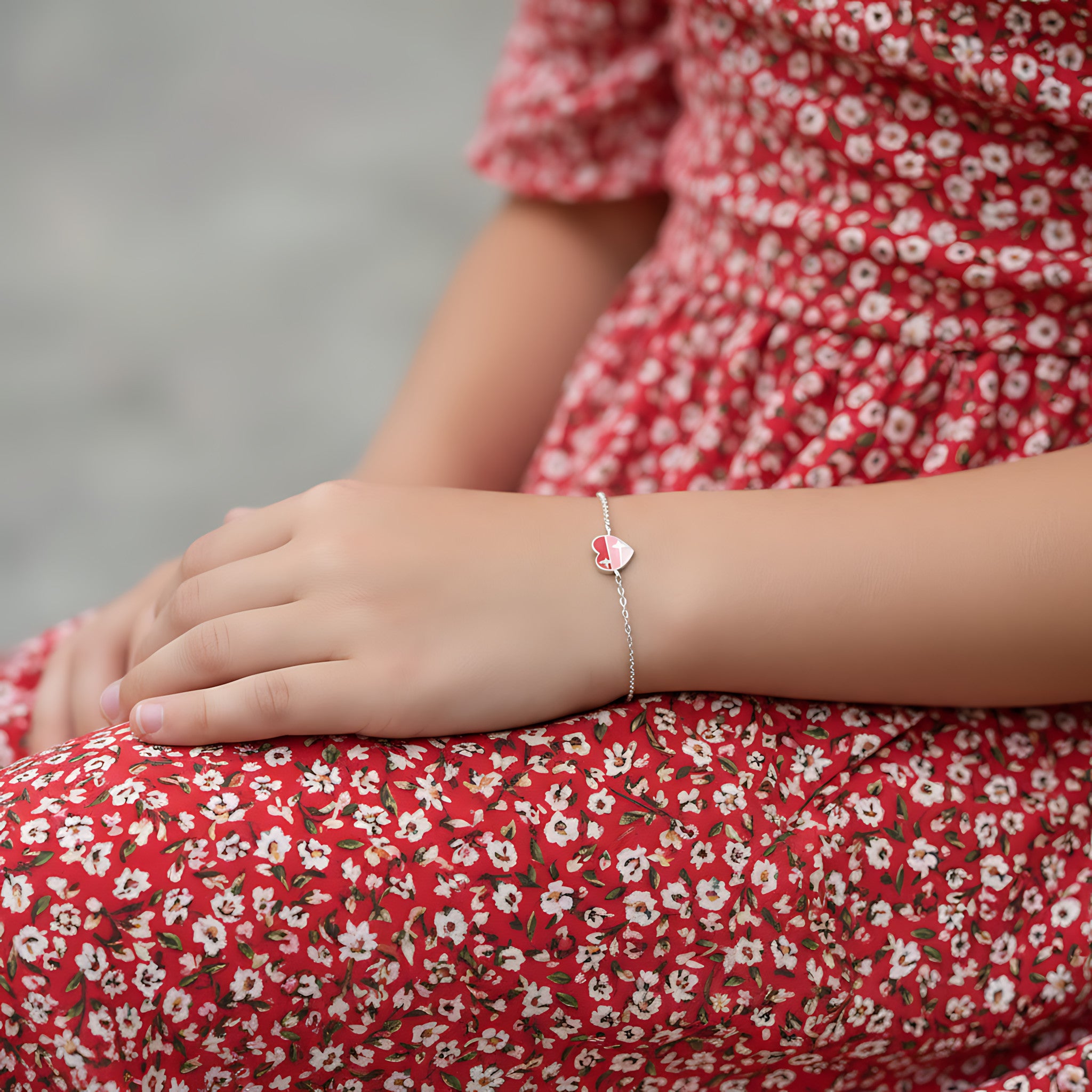 Silver bracelet with a red heart charm on a girl's wrist against a floral dress background