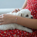 Closeup of a girl's arm wearing a silver heart bracelet and a red floral dress holding a small white dog