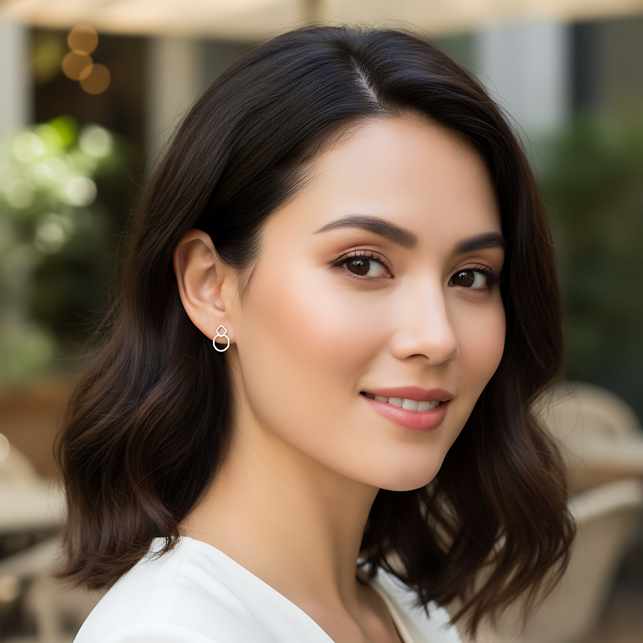 Woman with dark hair wearing stud earring  with a square and a circle intertwined and a white top in an outdoor setting