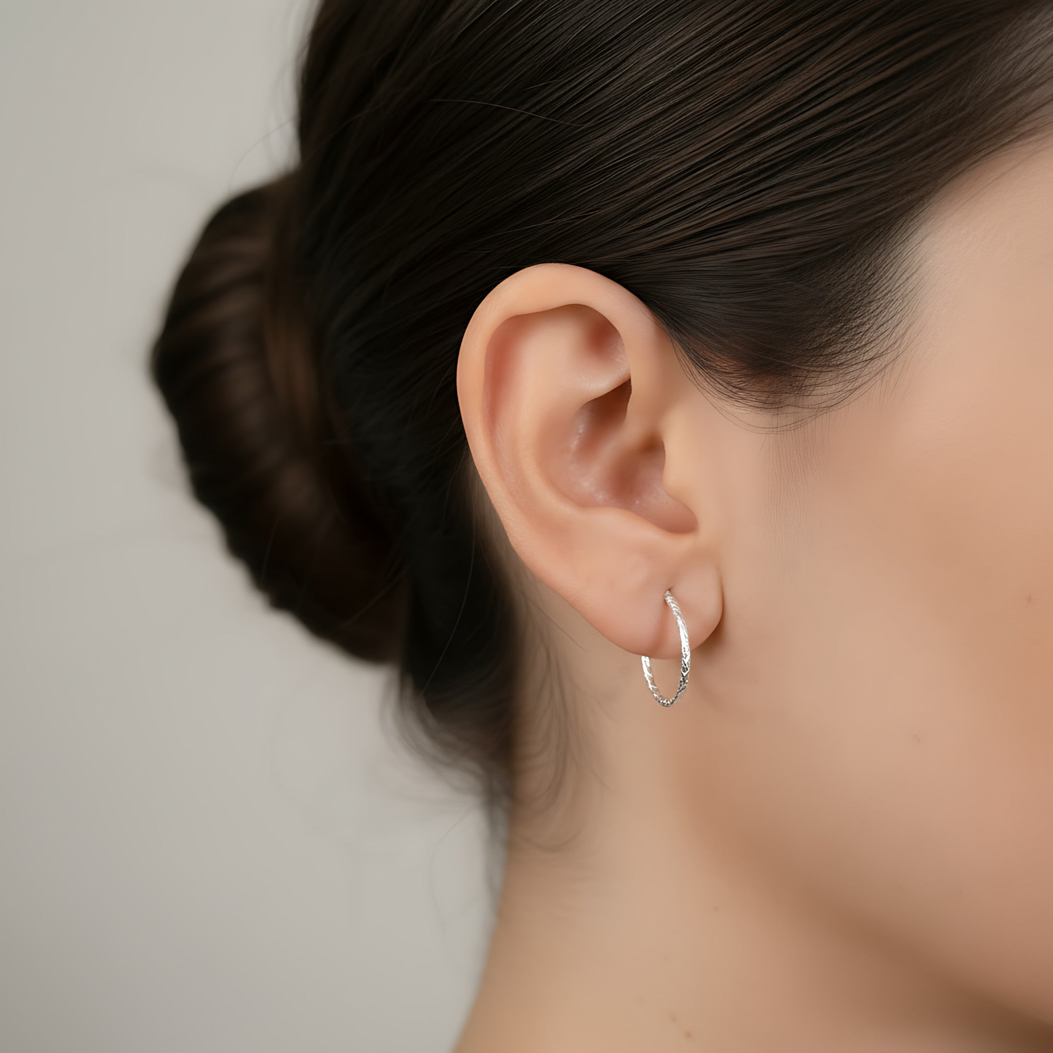 Close-up of an ear wearing an elongated silver textured half hoop stud earring with a neutral background